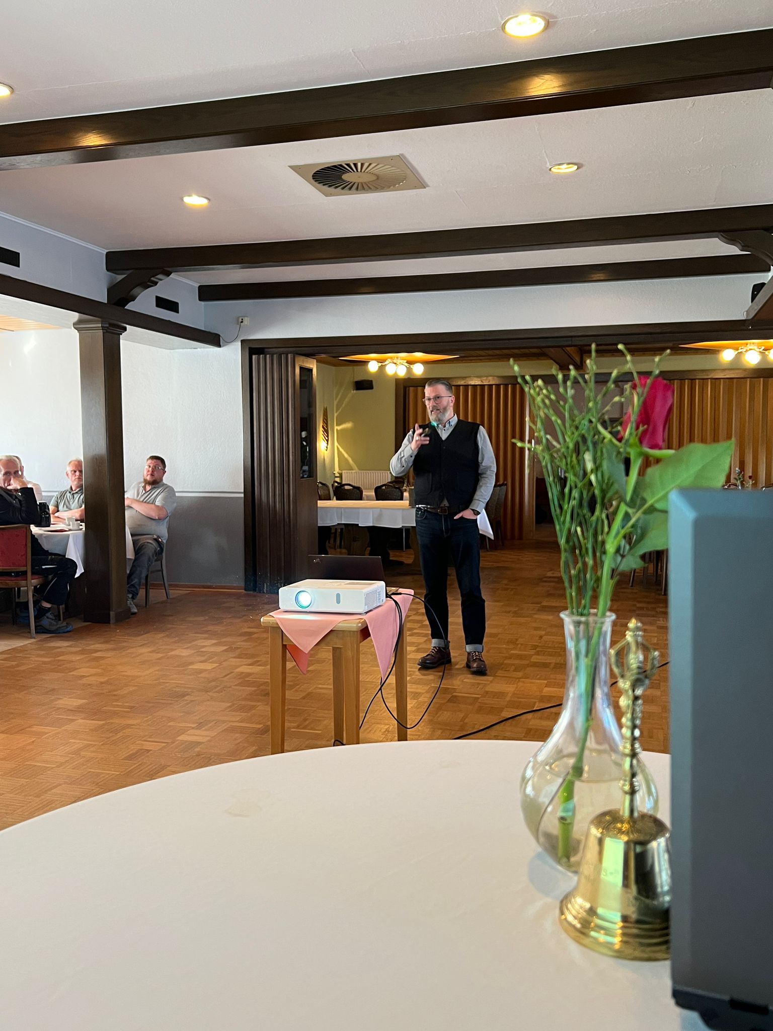Ein Mann steht in einem Saal und spricht, während er einen Zeigefinger hebt. Im Vordergrund befindet sich ein Tisch mit Blumen und einem Projektor auf einem kleinen Tisch. Im Hintergrund sitzen mehrere Personen mit Blick auf den Redner.