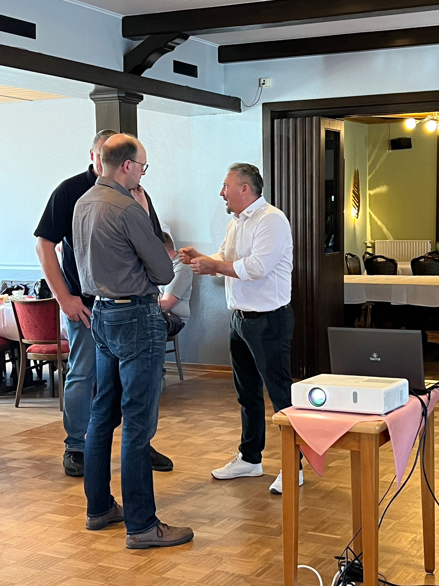 Eine Gruppe von vier Männern steht in einem Restaurant und diskutiert. Einer der Männer in einem weißen Hemd spricht lebhaft mit den anderen, während ein Projektor auf einem Tisch in der Nähe bereitsteht.
