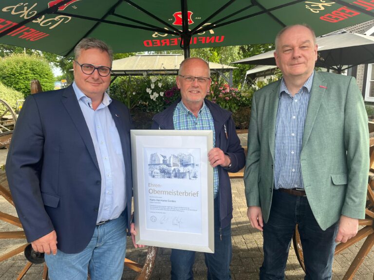 Drei Männer stehen nebeneinander im Freien unter einem großen Sonnenschirm; der Mann in der Mitte hält eine gerahmte Ehrenurkunde mit der Aufschrift "Ehren-Obermeisterbrief".