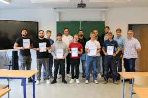 Gruppe von fünfzehn Männern in einem Klassenzimmer, von denen einige Urkunden in den Händen halten, lächeln und vor einer grünen Tafel posieren.