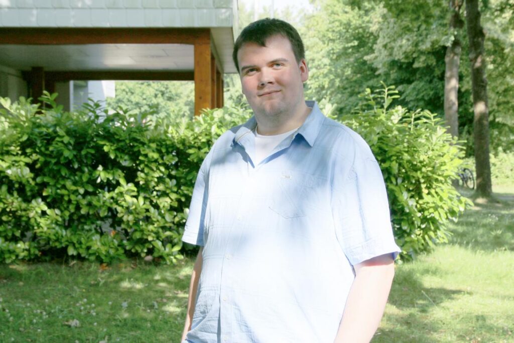 Mann mit hellblauem Hemd steht im Grünen vor einer Hecke, im Hintergrund sind Bäume und ein Gebäude zu sehen.