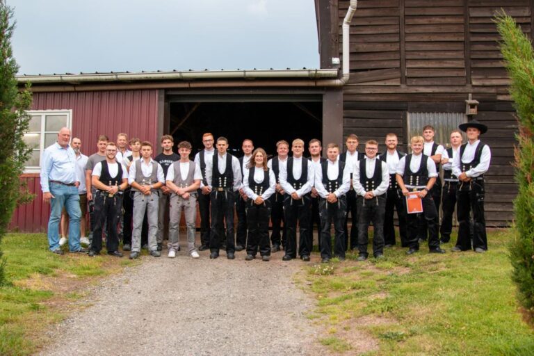 Gruppenfoto von etwa zwanzig jungen Männern und einer Frau vor einem Holz- und Backsteingebäude, viele davon in traditioneller Zunftkleidung der Zimmerer mit weißen Hemden und schwarzen Westen oder Jacken.