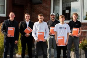 Sechs junge Männer stehen nebeneinander vor einem Gebäude und halten orangefarbene Mappen mit der Aufschrift "Willkommen bei den Profis" in den Händen, alle lächeln freundlich in die Kamera.