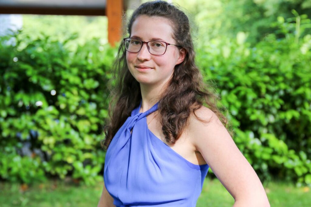 Junge Frau mit lockigen braunen Haaren und Brille posiert im Freien vor einer grünen Hecke, trägt ein ärmelloses, blaues Oberteil mit Knoten am Hals.