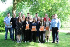 Gruppenfoto von 16 jungen Erwachsenen und zwei älteren Männern, teilweise mit Dokumenten in der Hand, im Freien bei sonnigem Wetter unter Bäumen.