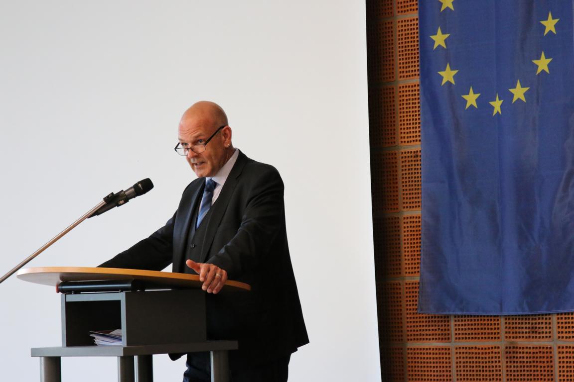 Mann im Anzug spricht an einem Rednerpult mit Mikrofon, daneben eine Europaflagge, vermutlich auf einer Konferenz oder bei einer offiziellen Veranstaltung.