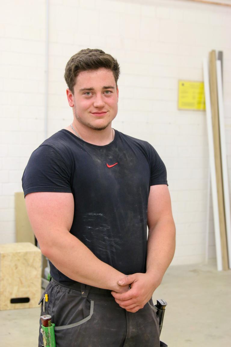 Junger, muskulöser Handwerker mit kurzem, dunklem Haar steht vor einer weißen Backsteinwand, trägt ein schwarzes, leicht verschmutztes T-Shirt und Werkzeuggürtel.