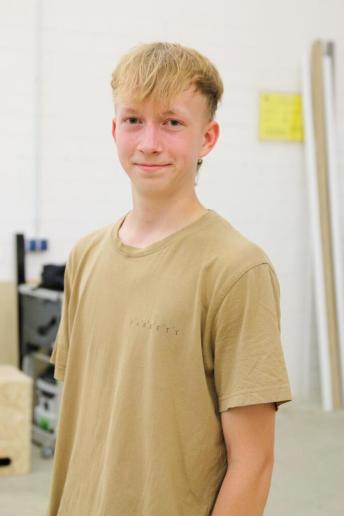 Junger Mann mit blonden Haaren und beigem T-Shirt steht in einem hellen Raum, wahrscheinlich eine Werkstatt, und lächelt in die Kamera.