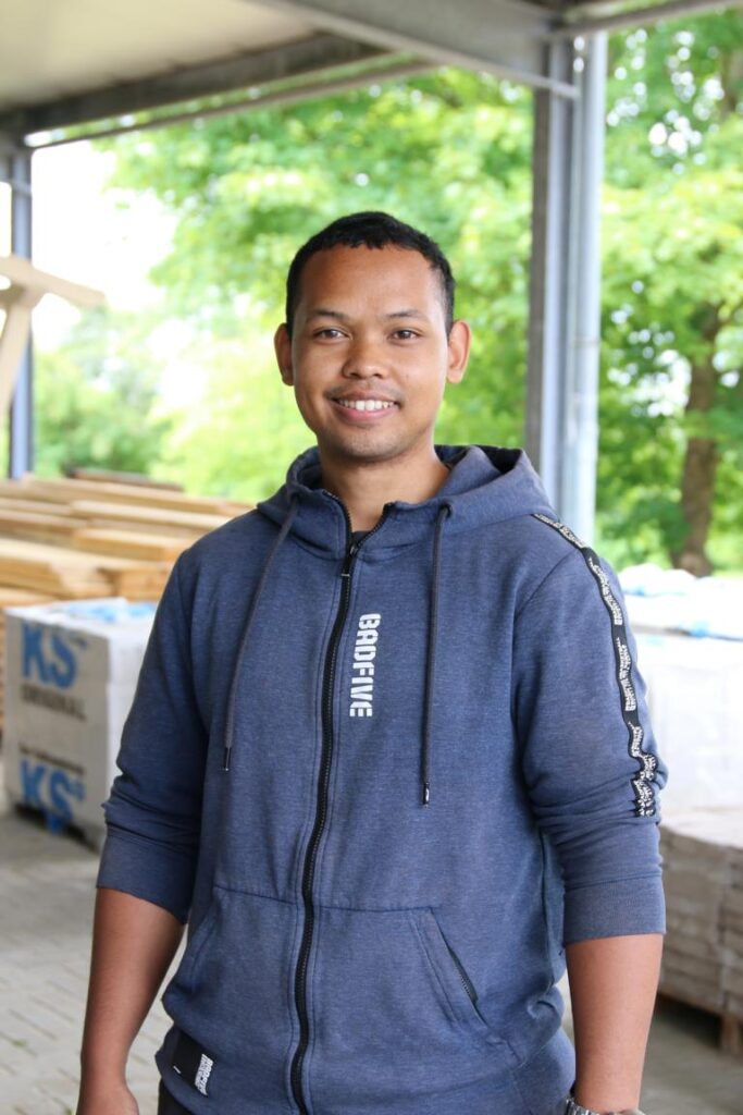 Mann mit dunklen kurzen Haaren und blauem Kapuzenpulli lächelt, im Hintergrund Lagerhalle mit Holzstapeln und grünem Baum draußen.