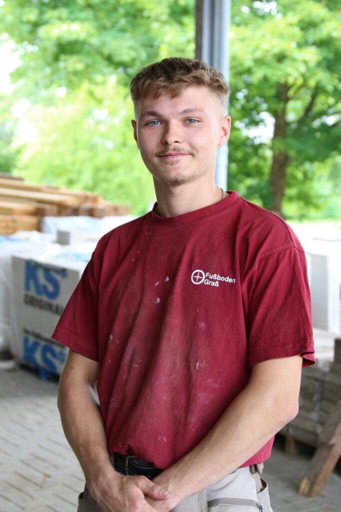 Junger Handwerker mit kurzem, blondem Haar trägt ein rotes, verschmutztes T-Shirt mit dem Aufdruck 'Fußboden Graß' und steht in einer Werkstattumgebung mit Verpackungen und Holzstapeln im Hintergrund.