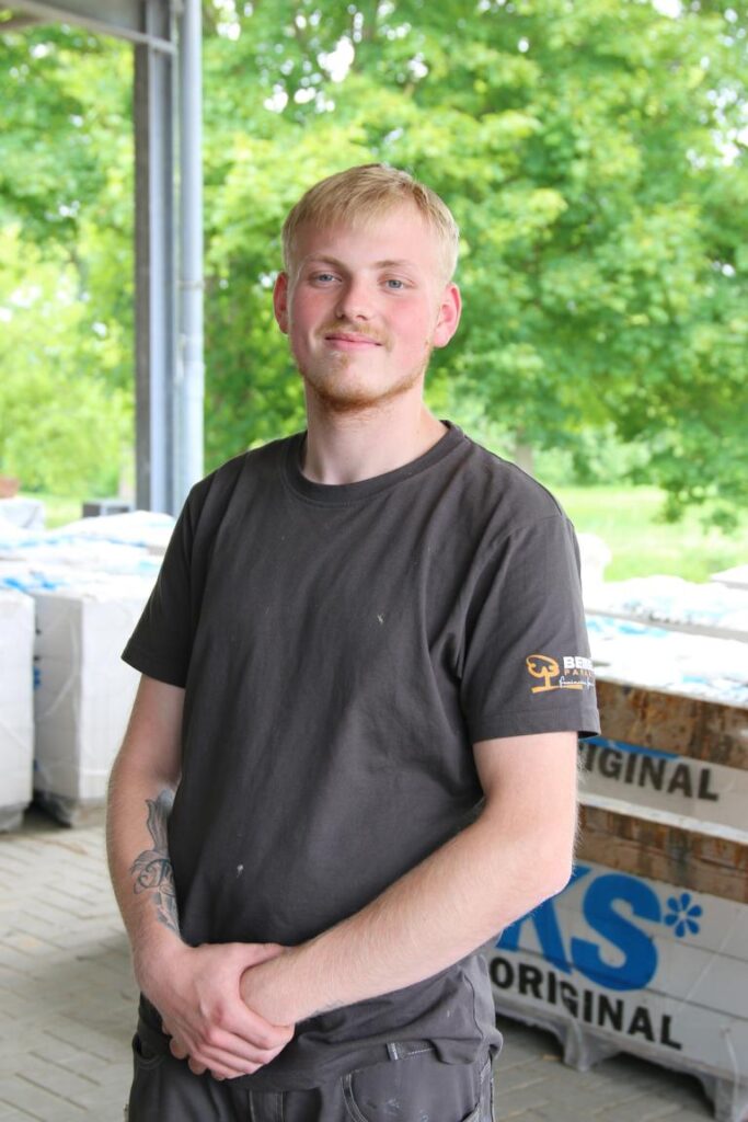 Junger Mann mit blondem Haar und kurzem Bart steht in Arbeitskleidung lächelnd vor Paletten im Freien, im Hintergrund grüne Bäume.