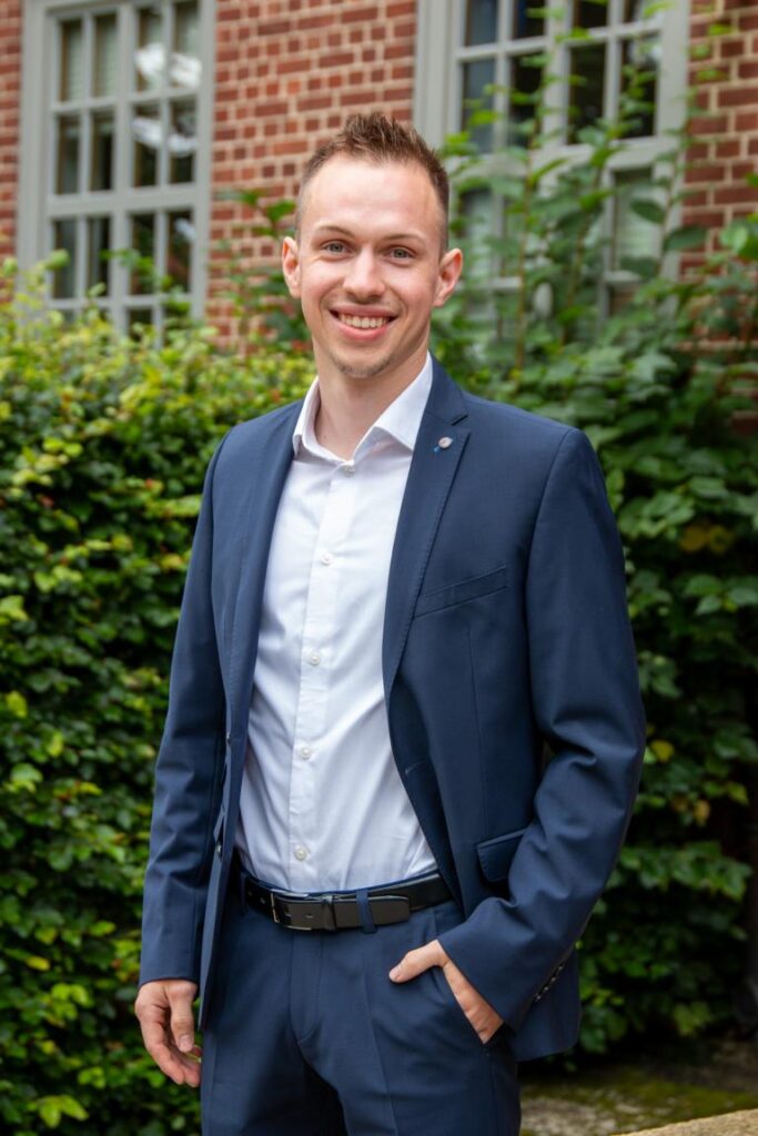 Junger Mann in einem dunkelblauen Anzug mit weißem Hemd, lächelt freundlich vor einer grünen Hecke und einem Backsteingebäude.