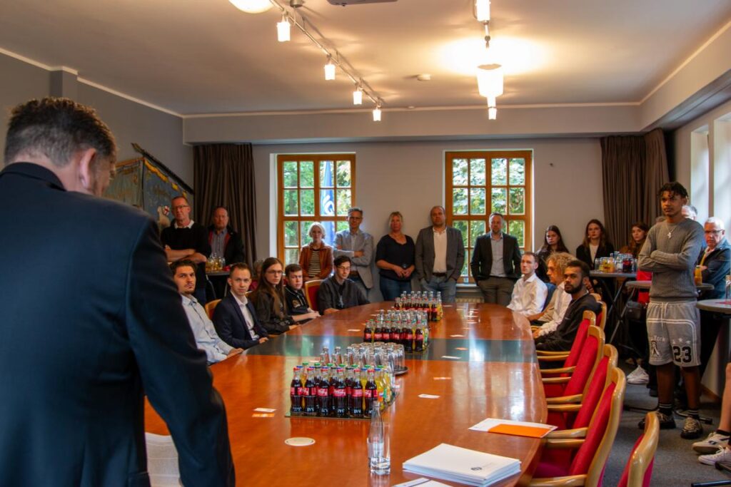 Besprechung in einem Konferenzraum mit Teilnehmern, die an einem langen Tisch sitzen und stehen. Auf dem Tisch stehen Getränke, darunter Flaschen mit Cola. Die Atmosphäre ist formell, mit einer Vielzahl von Menschen, die aufmerksam einer Präsentation folgen.