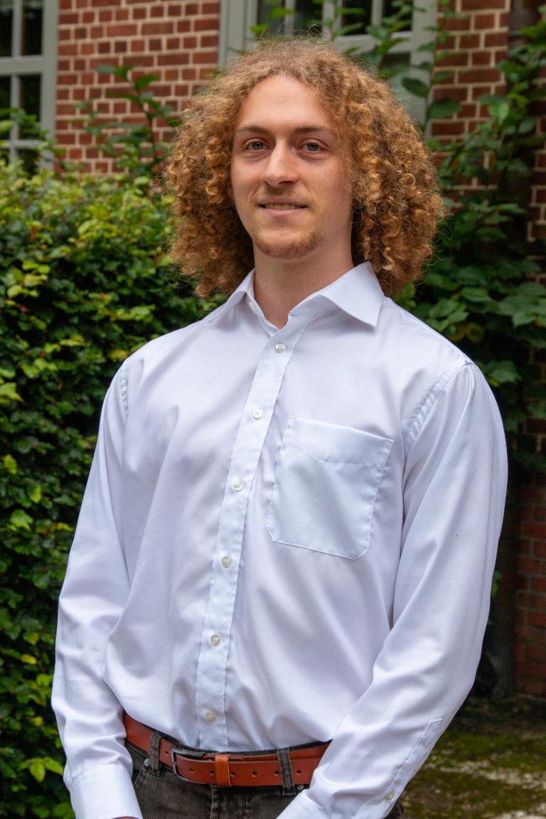 Junger Mann mit lockigem Haar in einem weißen Hemd steht vor einer grünen Hecke und einem Backsteingebäude.
