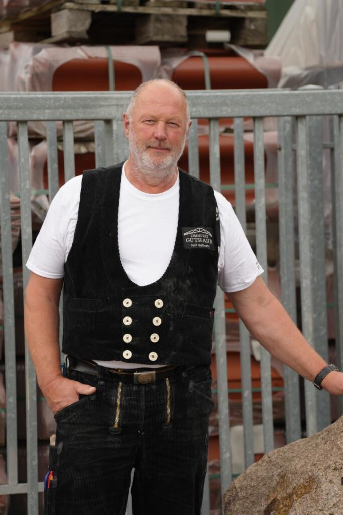 Ein Handwerker in traditioneller Kleidung steht vor einem Zaun, im Hintergrund sind Paletten mit Baustellenmaterialien sichtbar. Er trägt ein schwarzes Weste mit Knöpfen und hat einen Bart.