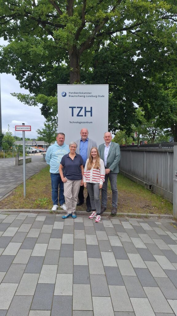 Gruppenfoto vor dem Technologiezentrum TZH der Handwerkskammer Braunschweig-Lüneburg-Stade, mit fünf Personen, die in der Mitte stehen, umgeben von Bäumen und einem gepflasterten Weg.
