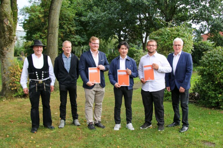 Gruppenfoto von sechs Personen im Freien, die stolz ihre Zertifikate mit der Aufschrift „Willkommen im neuen Profil“ präsentieren. Die Szene spielt in einem grünen Garten mit Bäumen im Hintergrund.