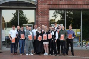 Gruppe von Absolventen hält Urkunden in den Händen, posiert vor einem Gebäude mit großen Fenstern. Sie tragen verschiedene formelle Outfits und lächeln in die Kamera. Die Szene vermittelt Freude und Feierlichkeit im Rahmen einer Abschlusszeremonie.