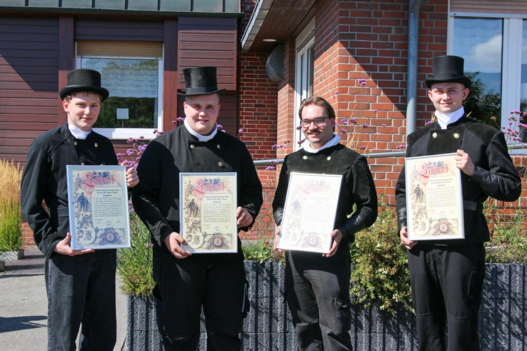 Vier Männer in traditionellen schwarzen Anzügen und Zylinder halten Urkunden in der Hand. Sie stehen vor einem Backsteingebäude, umgeben von Blumen. Die Urkunden scheinen besondere Auszeichnungen oder Zertifikate zu sein.