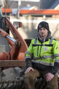 Junger Bauarbeiter in leuchtend gelber Sicherheitsjacke sitzt auf einem Stapel Steine, im Hintergrund ist ein Bagger sichtbar.