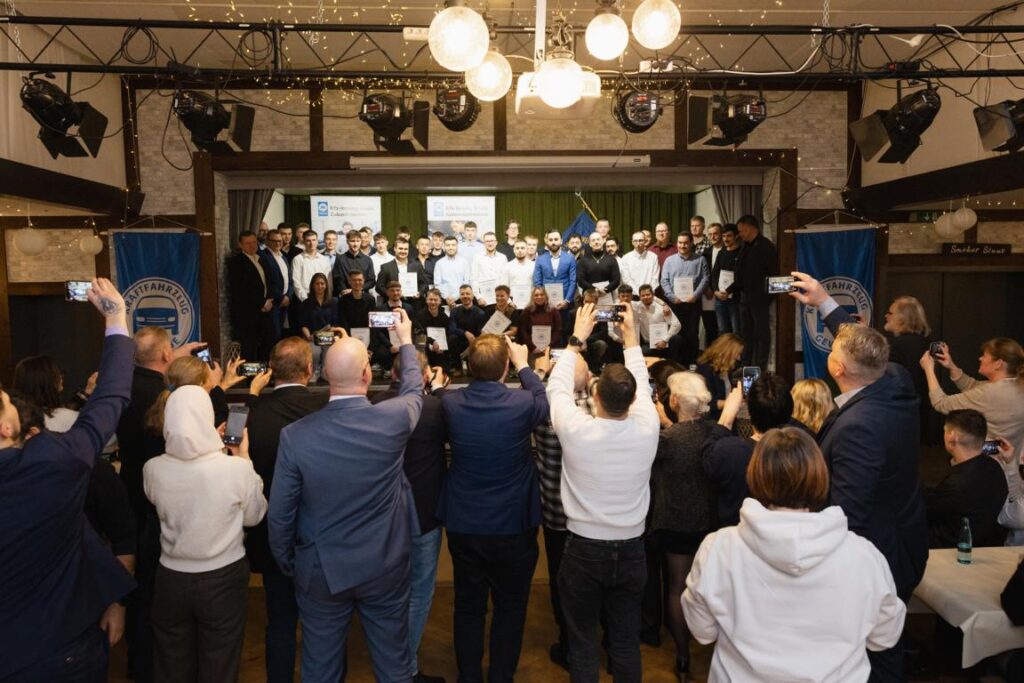 Gruppenfoto von Teilnehmern einer Preisverleihung in einem festlich dekorierten Saal, umgeben von Zuschauern, die mit Smartphones die Veranstaltung festhalten. Die Teilnehmer halten Urkunden in den Händen und stehen vor einer Bannerwand mit dem Logo des Veranstalters.