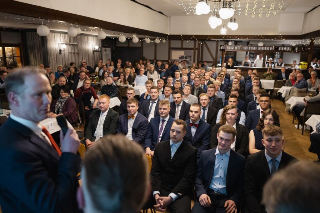 Versammlung von Gästen in formeller Kleidung bei einer Veranstaltung, während ein Redner im Vordergrund spricht. Die Szene zeigt eine große Menge, die aufmerksam zuhört, in einem festlich dekorierten Raum mit Tischen.