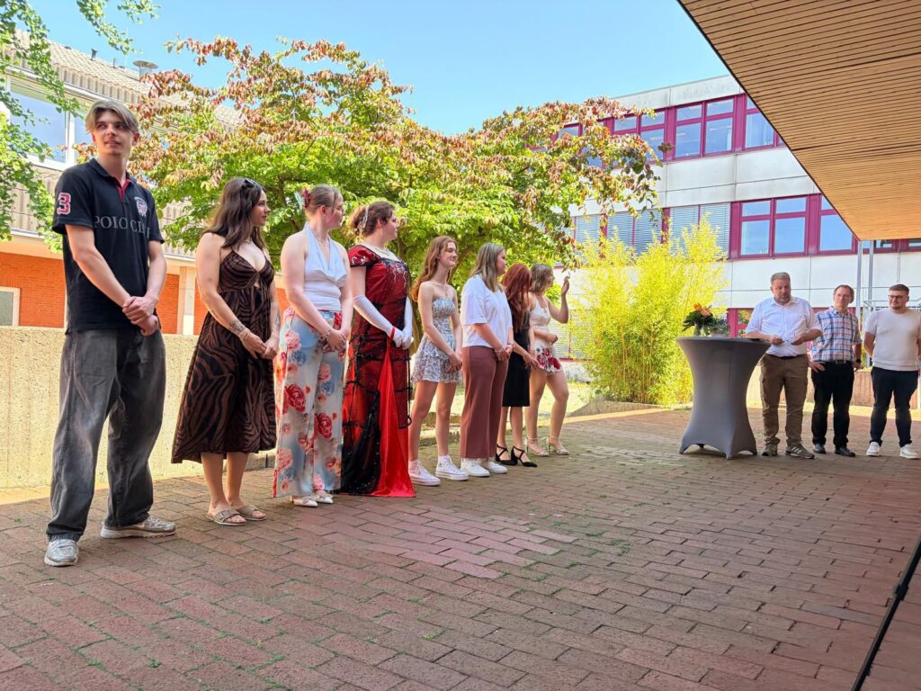 Gruppe von Personen in festlicher Kleidung steht im Freien bei einer Veranstaltung, während ein Redner an einem Tisch spricht. Im Hintergrund sind Bäume und ein modernes Gebäude sichtbar.
