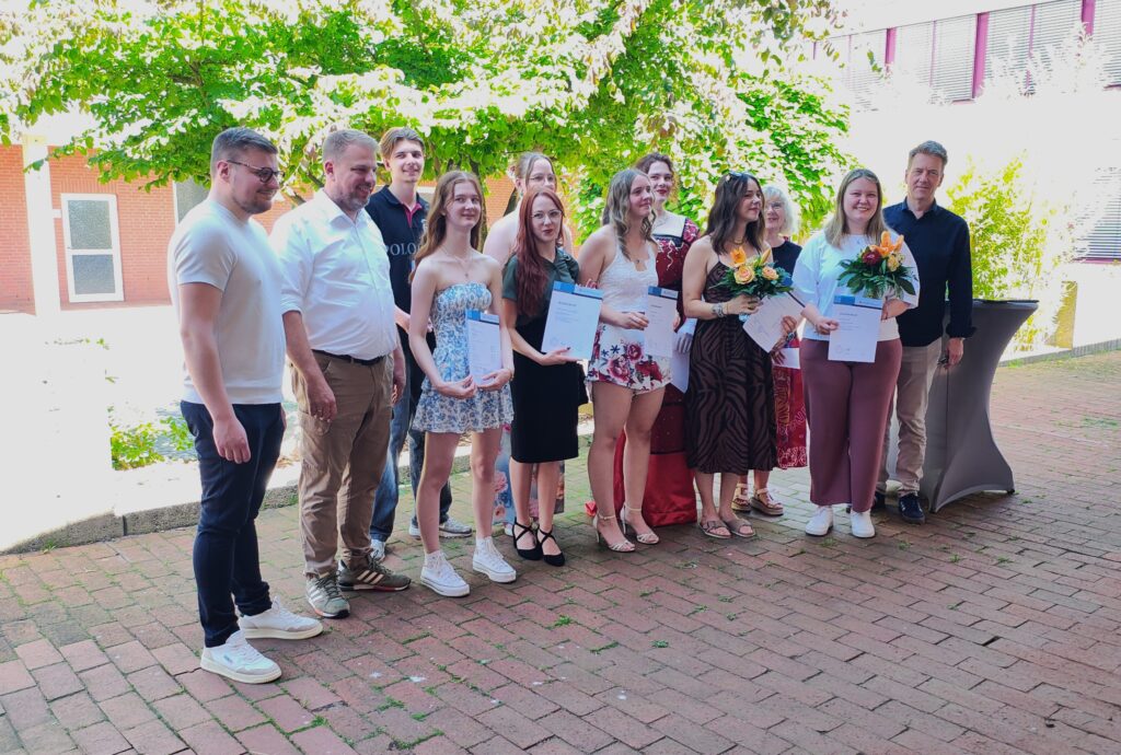 Gruppe von Absolventen feiert ihre Abschlusszeremonie im Freien, hält Urkunden und Blumen in der Hand. Sie stehen zusammen in einer Reihe, umgeben von grüner Vegetation und einem festlichen Ambiente.