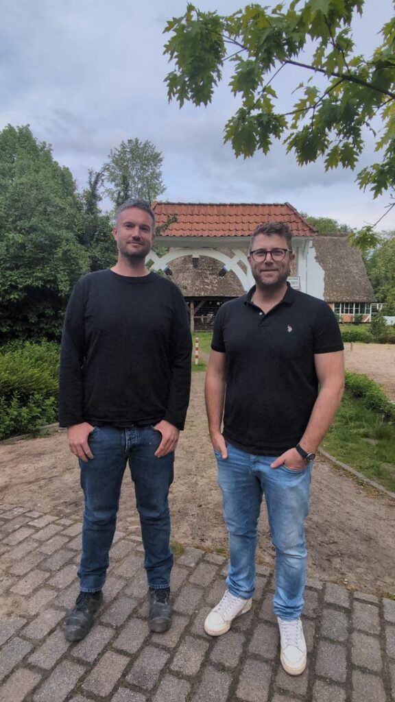 Zwei Männer stehen auf einem gepflasterten Weg vor einem traditionellen, reetgedeckten Gebäude. Der Mann links trägt ein schwarzes Sweatshirt und Jeans, während der Mann rechts ein schwarzes Poloshirt und helle Jeans trägt. Im Hintergrund sind Bäume und eine gepflegte Landschaft sichtbar.