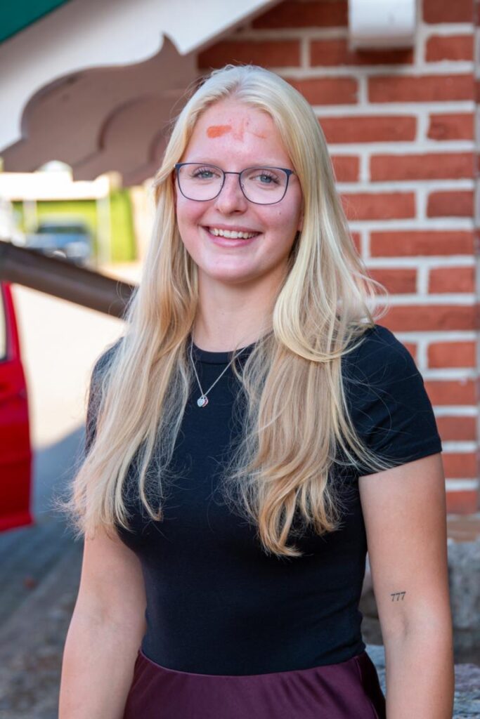 Junge blonde Frau mit Brille und schwarzem T-Shirt steht lächelnd vor einer Backsteinwand, rotes Auto im Hintergrund, Tattoo 777 am Arm, roter Fleck auf der Stirn.