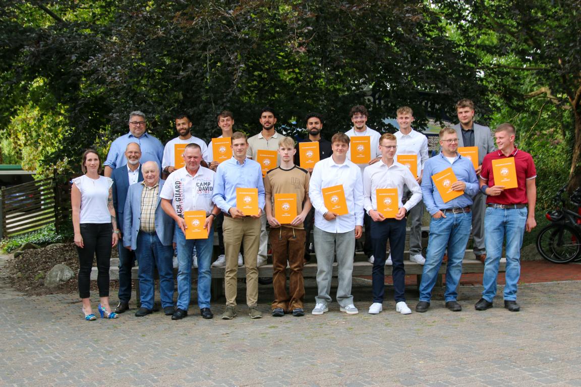 Gruppe von Absolventen, die stolz ihre Zertifikate in den Händen halten, fotografiert im Freien vor einer grünen Kulisse. Die Männer und Frauen, angezogen in formeller Kleidung, stehen auf einer Bank und lächeln in die Kamera.