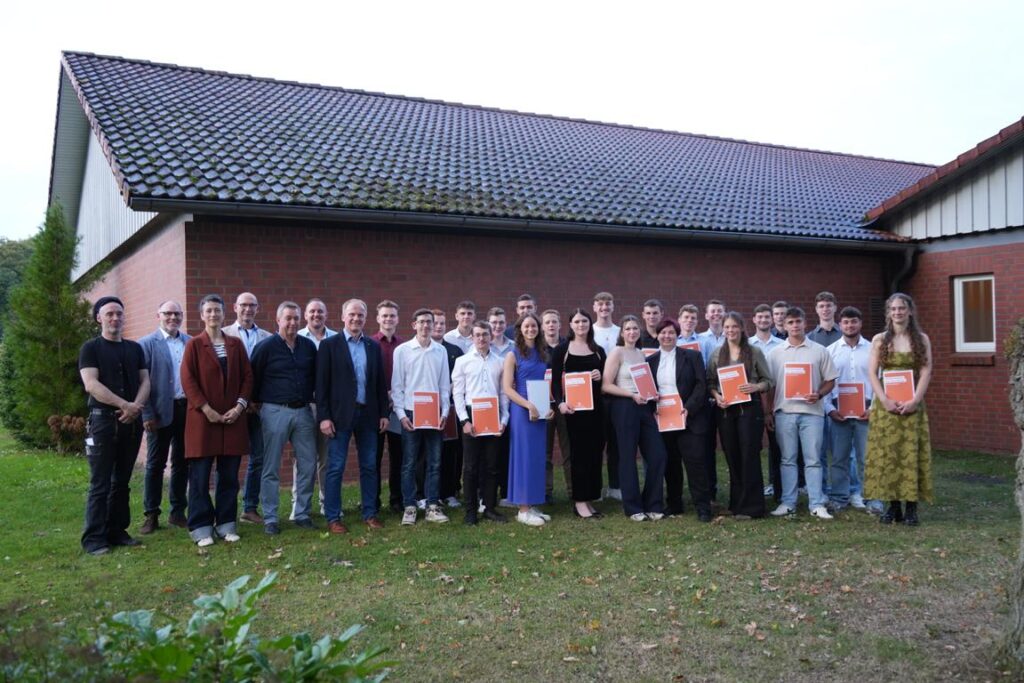 Gruppenfoto von jungen Erwachsenen und Ausbildern bei einer Abschlussfeier vor einem Backsteingebäude, mehrere Personen halten Urkunden in der Hand