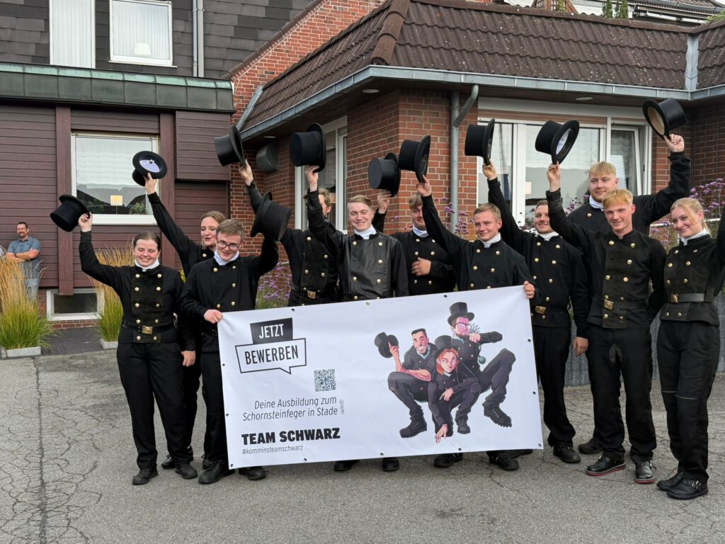 Elf frisch ausgebildete Schornsteinfegerinnen und Schornsteinfeger in traditioneller Arbeitskleidung feiern ihren Abschluss vor einem Gebäude und halten einen Banner mit Ausbildungsinformation hoch.