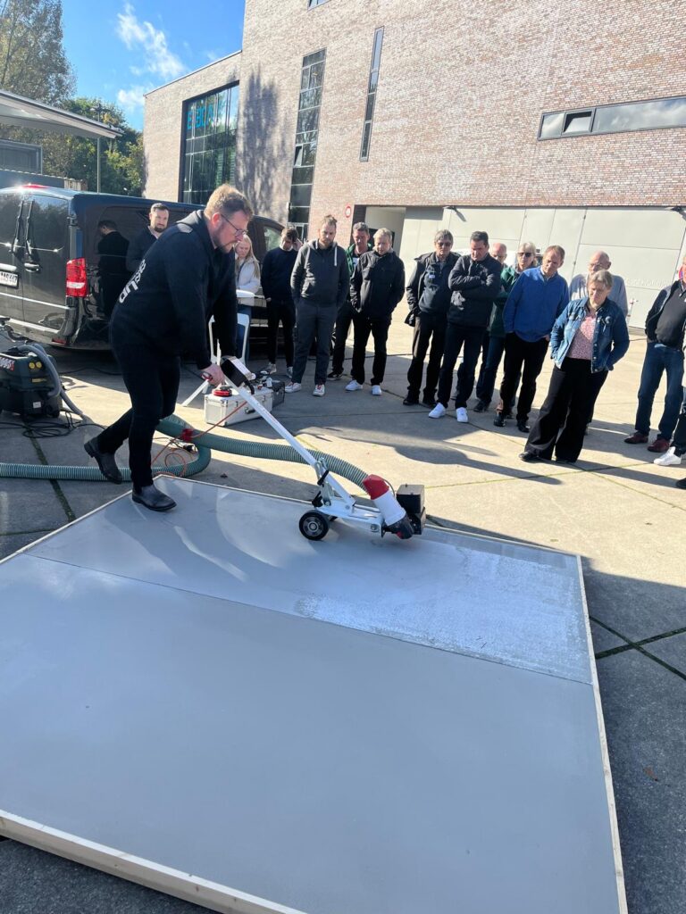 Mann demonstriert Bodenschleifmaschine auf Betonfläche vor einer Gruppe von Zuschauern im Freien bei einer Produktschulung